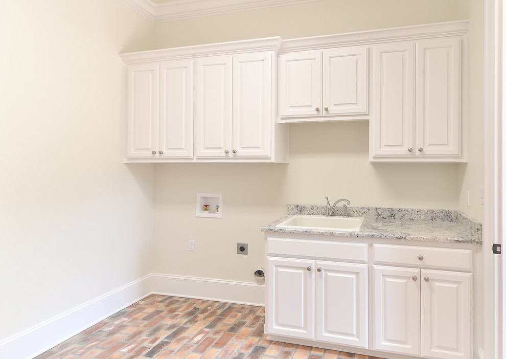Photo of a large traditional dedicated laundry room in New Orleans with a drop-in sink, raised-panel cabinets, white cabinets, granite benchtops, beige walls, brick floors and a side-by-side washer and dryer.