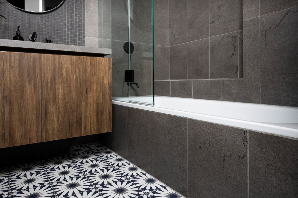 Photo of a small industrial master bathroom in Melbourne with flat-panel cabinets, dark wood cabinets, a drop-in tub, a shower/bathtub combo, a one-piece toilet, gray tile, porcelain tile, grey walls, ceramic floors, an undermount sink, engineered quartz benchtops, multi-coloured floor, an open shower, grey benchtops, a shower seat, a single vanity and a floating vanity.