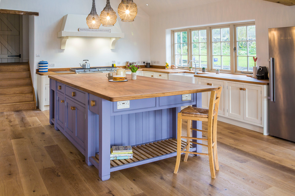 This is an example of a country l-shaped kitchen in Other with a farmhouse sink, shaker cabinets, white cabinets, wood benchtops, stainless steel appliances, light hardwood floors and with island.