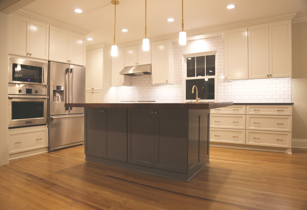 Example of a mid-sized l-shaped light wood floor eat-in kitchen design with a farmhouse sink, recessed-panel cabinets, white cabinets, granite countertops, white backsplash, ceramic backsplash, stainless steel appliances and an island