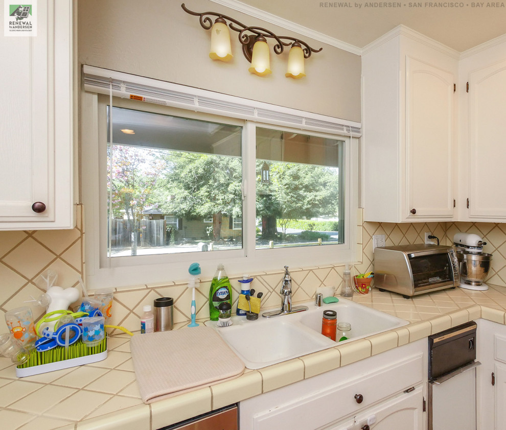 This is an example of a mid-sized u-shaped eat-in kitchen in San Francisco with a drop-in sink, white cabinets, tile benchtops, beige splashback, ceramic splashback, stainless steel appliances, beige benchtop and vaulted.