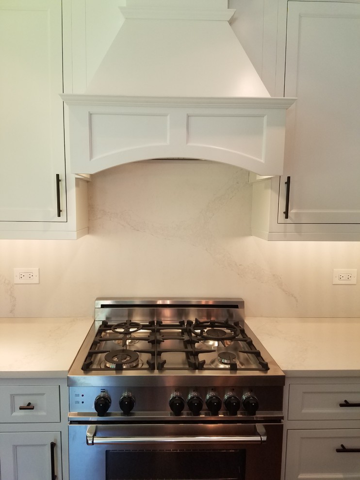 Mid-sized transitional l-shaped medium tone wood floor and brown floor eat-in kitchen photo in Chicago with a farmhouse sink, recessed-panel cabinets, white cabinets, quartz countertops, white backsplash, stainless steel appliances and an island