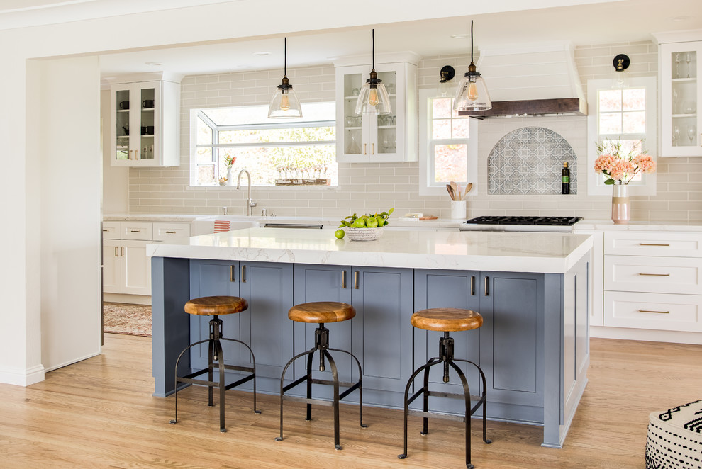 Design ideas for a country galley kitchen in San Francisco with a farmhouse sink, shaker cabinets, white cabinets, grey splashback, stainless steel appliances, light hardwood floors, with island and white benchtop.