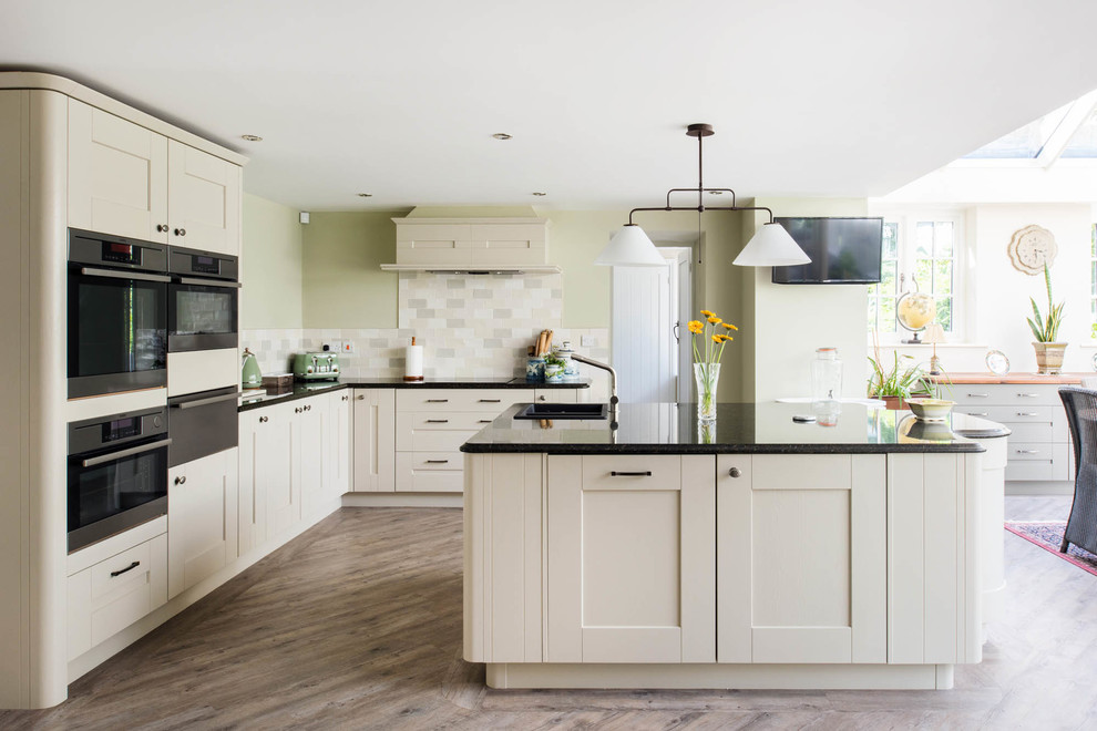 This is an example of a transitional l-shaped eat-in kitchen in Other with a drop-in sink, shaker cabinets, beige cabinets, multi-coloured splashback, mosaic tile splashback, black appliances, light hardwood floors and with island.