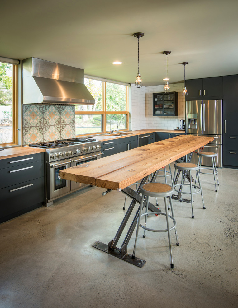 Photo of a mid-sized industrial l-shaped eat-in kitchen in Philadelphia with an undermount sink, flat-panel cabinets, black cabinets, wood benchtops, white splashback, subway tile splashback, stainless steel appliances, concrete floors and with island.