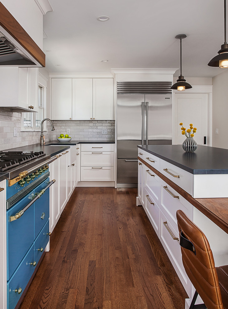 This is an example of a mid-sized transitional l-shaped eat-in kitchen in Detroit with a farmhouse sink, flat-panel cabinets, white cabinets, quartz benchtops, white splashback, ceramic splashback, stainless steel appliances, medium hardwood floors, with island, brown floor and black benchtop.