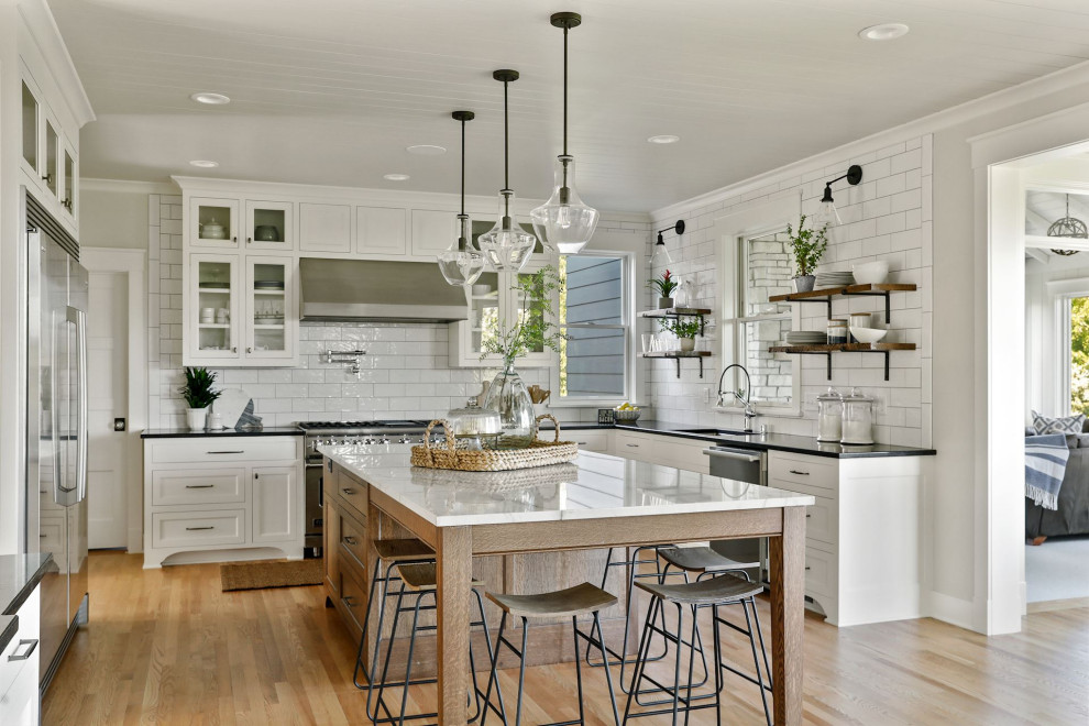 Design ideas for a country u-shaped kitchen in Minneapolis with an undermount sink, white cabinets, quartzite benchtops, subway tile splashback, stainless steel appliances, light hardwood floors, with island, shaker cabinets, white splashback, beige floor, black benchtop and timber.