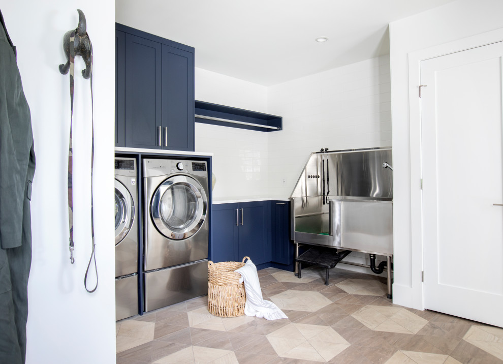 Photo of a large beach style l-shaped utility room in Vancouver with recessed-panel cabinets, blue cabinets, white splashback, porcelain splashback, white walls, porcelain floors, a side-by-side washer and dryer, brown floor and white benchtop.