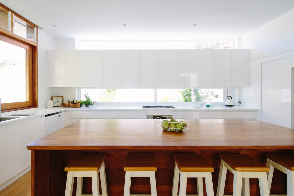 This is an example of a mid-sized contemporary l-shaped open plan kitchen in Sydney with an undermount sink, flat-panel cabinets, white cabinets, wood benchtops, window splashback, light hardwood floors and with island.