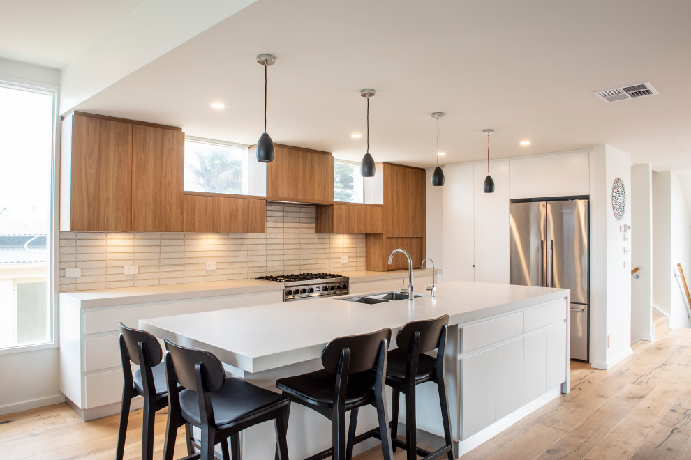 Large contemporary l-shaped open plan kitchen in Melbourne with an undermount sink, flat-panel cabinets, white cabinets, concrete benchtops, beige splashback, ceramic splashback, stainless steel appliances, medium hardwood floors, with island, grey benchtop and brown floor.