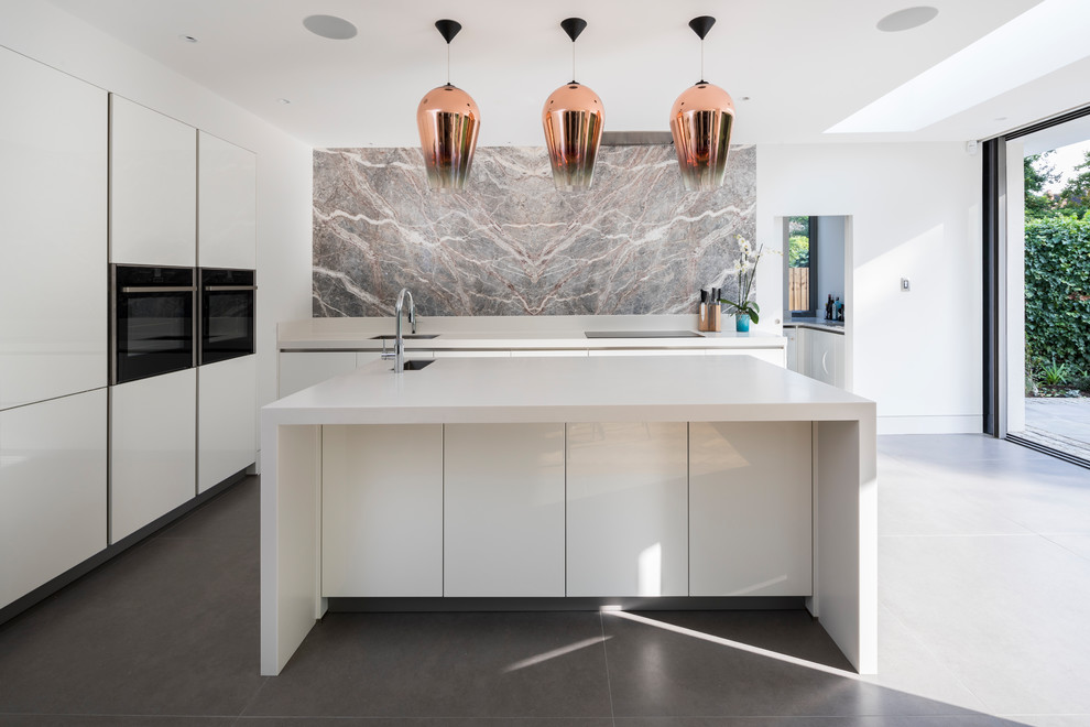 This is an example of a contemporary l-shaped kitchen in London with flat-panel cabinets, white cabinets, grey splashback, black appliances, with island, grey floor and white benchtop.