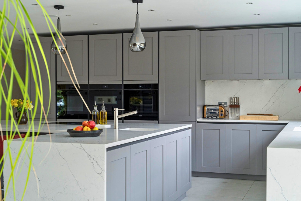 This is an example of a mid-sized transitional l-shaped open plan kitchen in Oxfordshire with a single-bowl sink, recessed-panel cabinets, grey cabinets, marble benchtops, black appliances, with island, grey floor and white benchtop.
