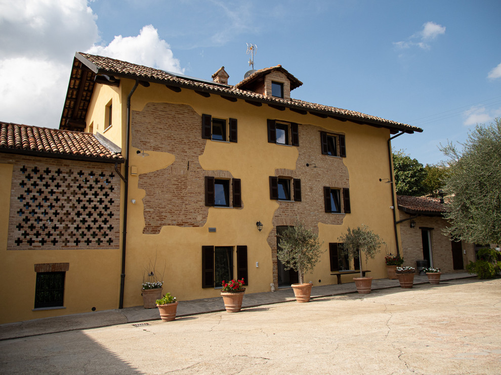 Esempio della villa ampia gialla country a tre piani con rivestimento in mattoni, tetto a capanna, tetto marrone e terreno in pendenza