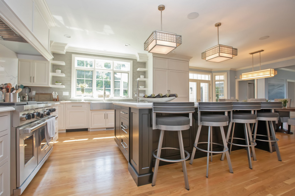 This is an example of a mid-sized modern u-shaped eat-in kitchen in Boston with a farmhouse sink, beaded inset cabinets, white cabinets, quartz benchtops, grey splashback, glass tile splashback, stainless steel appliances, light hardwood floors, with island, brown floor and white benchtop.