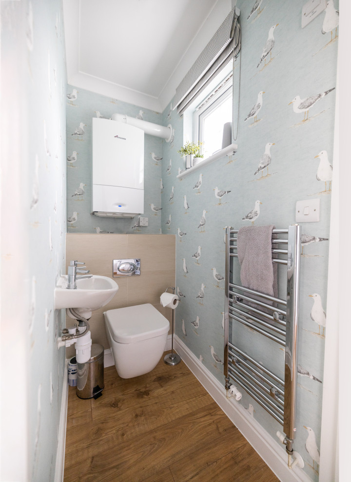 Small scandi cloakroom in Cornwall with a wall mounted toilet, beige tiles, ceramic tiles, blue walls, medium hardwood flooring, a wall-mounted sink, beige floors, a feature wall, a floating vanity unit and wallpapered walls.
