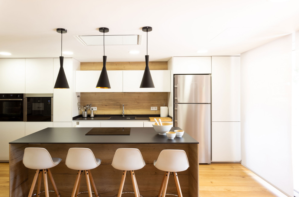 This is an example of a contemporary eat-in kitchen in Other with a drop-in sink, flat-panel cabinets, white cabinets, brown splashback, timber splashback, stainless steel appliances, medium hardwood floors, with island, brown floor and black benchtop.