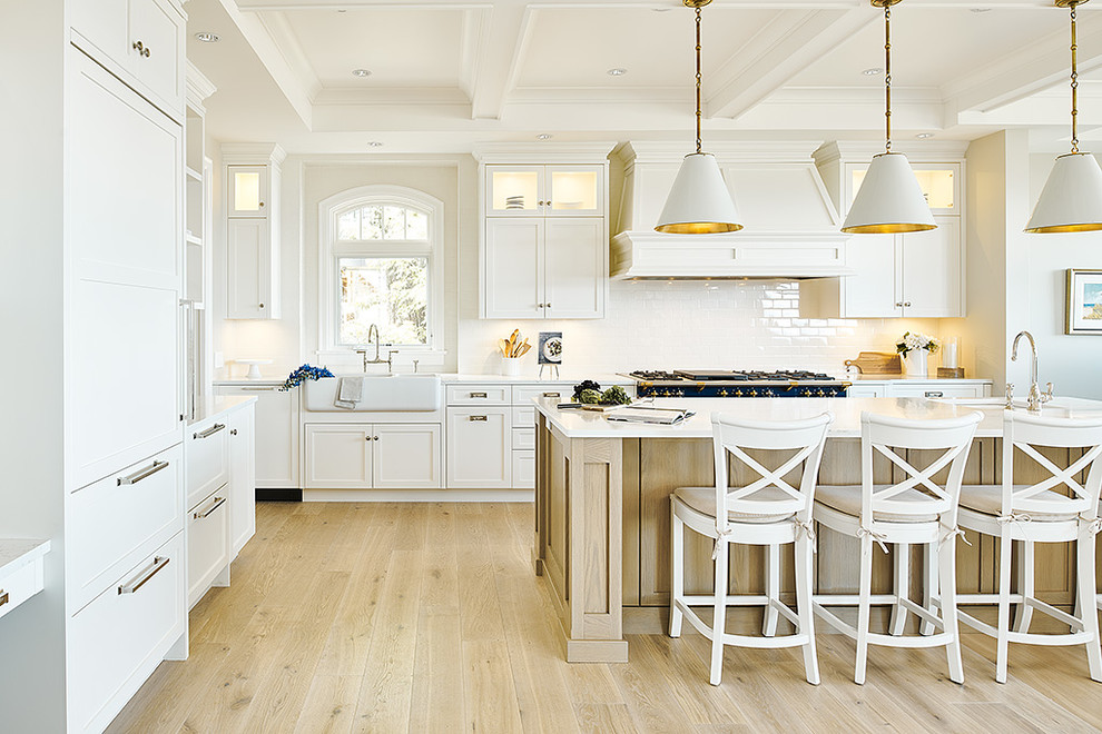 Photo of a large beach style l-shaped eat-in kitchen in Vancouver with a farmhouse sink, shaker cabinets, white cabinets, granite benchtops, white splashback, ceramic splashback, coloured appliances, light hardwood floors, with island, beige floor and white benchtop.