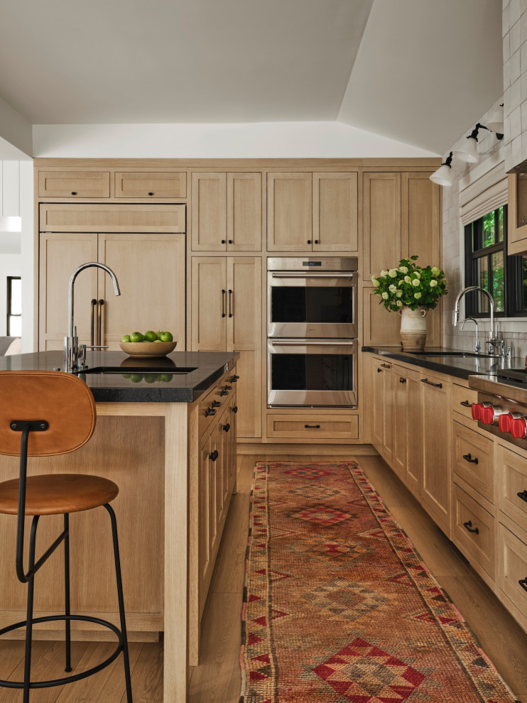 Design ideas for a transitional l-shaped kitchen in Los Angeles with an undermount sink, shaker cabinets, medium wood cabinets, white splashback, stainless steel appliances, medium hardwood floors, with island, brown floor and black benchtop.