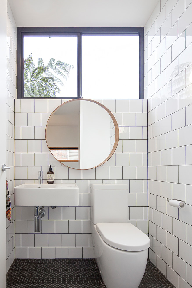 Photo of a midcentury powder room in Melbourne with white tile, white walls, mosaic tile floors, a wall-mount sink and black floor.