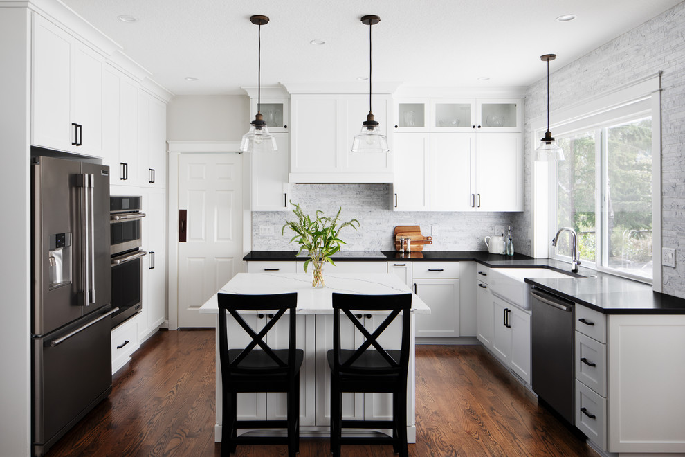 This is an example of a mid-sized contemporary u-shaped eat-in kitchen in Portland with a farmhouse sink, shaker cabinets, white cabinets, granite benchtops, white splashback, marble splashback, stainless steel appliances, dark hardwood floors, with island, brown floor and black benchtop.