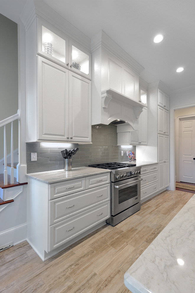 Large contemporary l-shaped open plan kitchen in Charleston with a farmhouse sink, raised-panel cabinets, white cabinets, granite benchtops, grey splashback, glass tile splashback, stainless steel appliances, porcelain floors and with island.