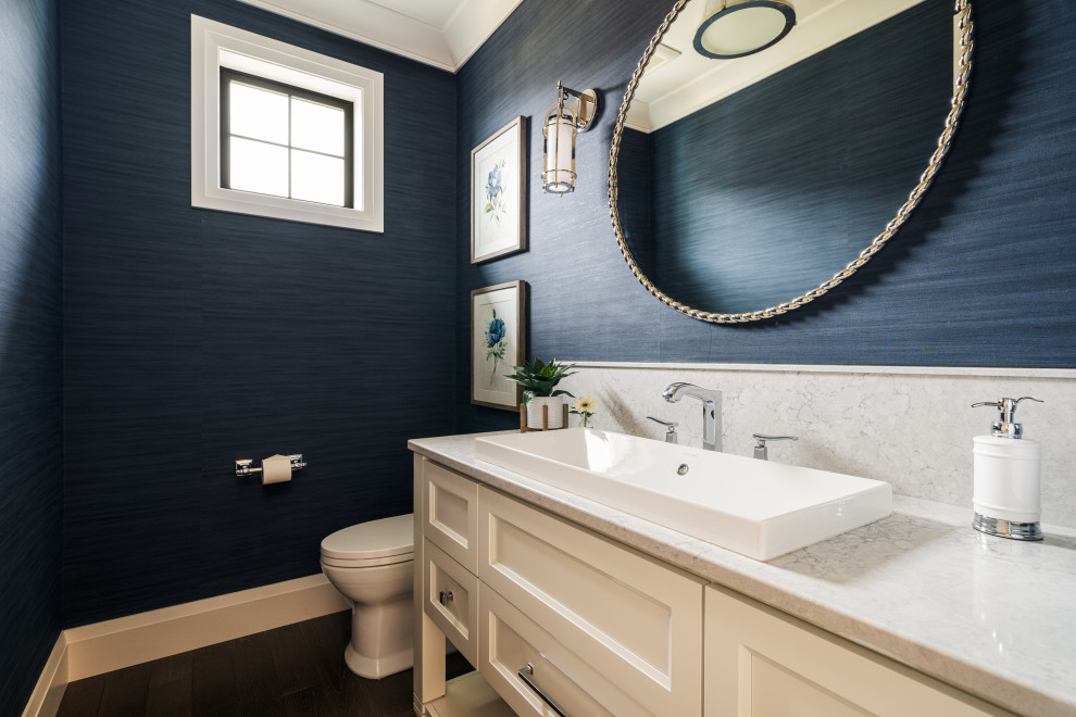 Medium sized beach style cloakroom in Seattle with recessed-panel cabinets, white cabinets, a one-piece toilet, blue walls, medium hardwood flooring, a vessel sink, engineered stone worktops, brown floors, grey worktops, a built in vanity unit and wallpapered walls.