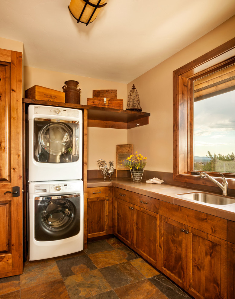 This is an example of a mid-sized country l-shaped dedicated laundry room in Other with an undermount sink, recessed-panel cabinets, dark wood cabinets, tile benchtops, beige walls, slate floors, a stacked washer and dryer, brown floor and beige benchtop.