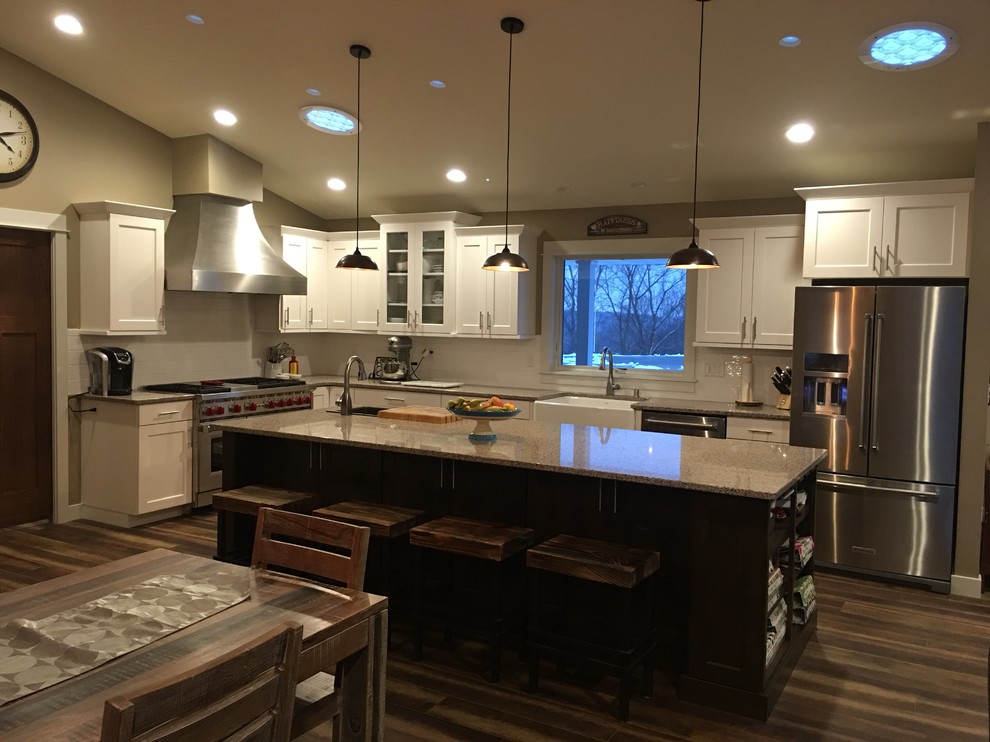 Large transitional l-shaped dark wood floor and brown floor eat-in kitchen photo with a farmhouse sink, shaker cabinets, white cabinets, granite countertops, white backsplash, stainless steel appliances and an island