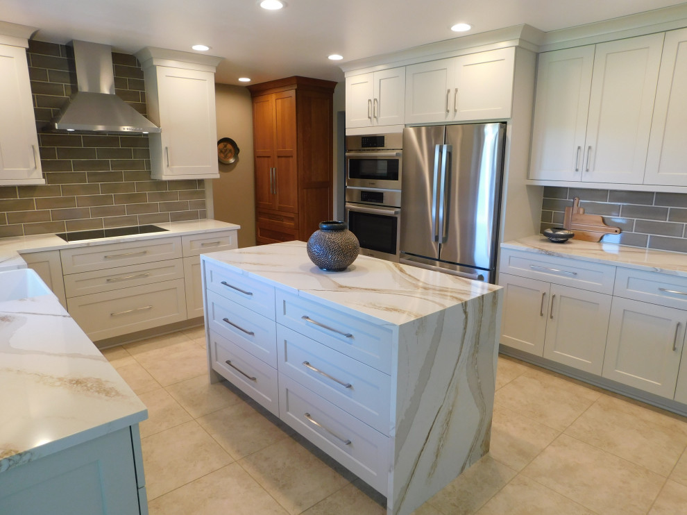Example of a transitional l-shaped porcelain tile and beige floor eat-in kitchen design with a farmhouse sink, flat-panel cabinets, quartz countertops, gray backsplash, porcelain backsplash, stainless steel appliances and an island