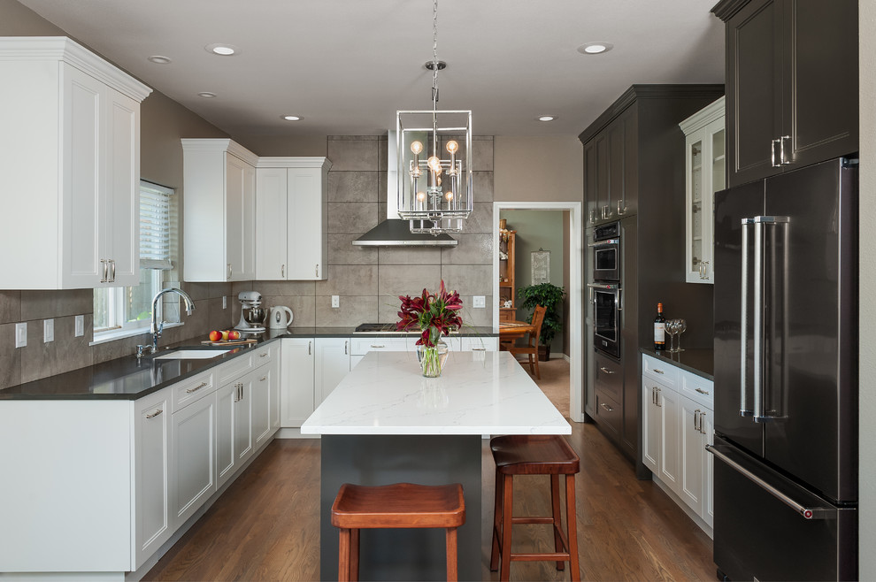 This is an example of a mid-sized transitional u-shaped open plan kitchen in Portland with a single-bowl sink, recessed-panel cabinets, white cabinets, quartz benchtops, grey splashback, porcelain splashback, stainless steel appliances, medium hardwood floors, with island, grey floor and white benchtop.