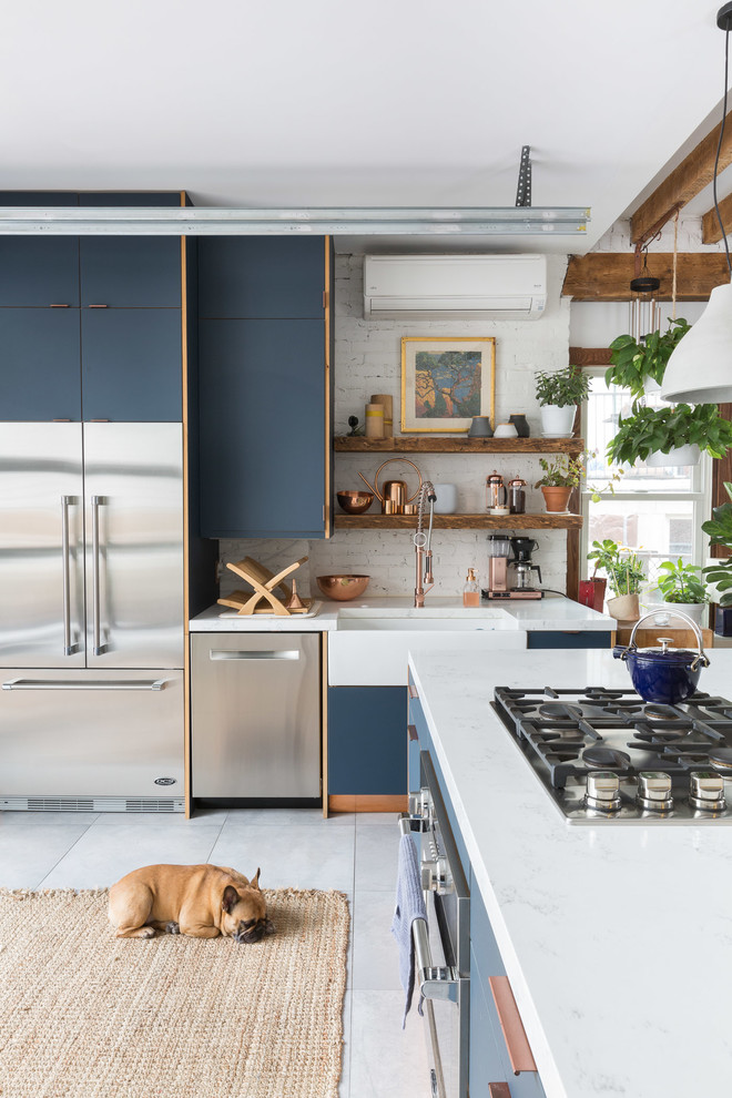 Eclectic l-shaped kitchen in New York with a farmhouse sink, flat-panel cabinets, blue cabinets, white splashback, brick splashback, stainless steel appliances, with island, grey floor and white benchtop.