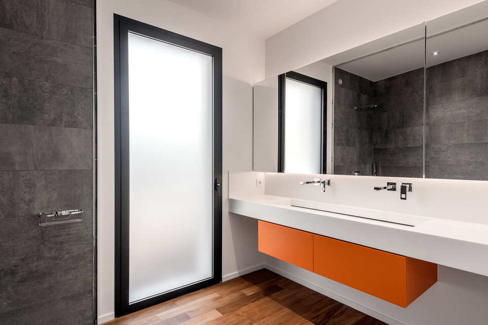 This is an example of a contemporary bathroom in Bordeaux with flat-panel cabinets, orange cabinets, gray tile, white walls, medium hardwood floors, a trough sink and white benchtops.