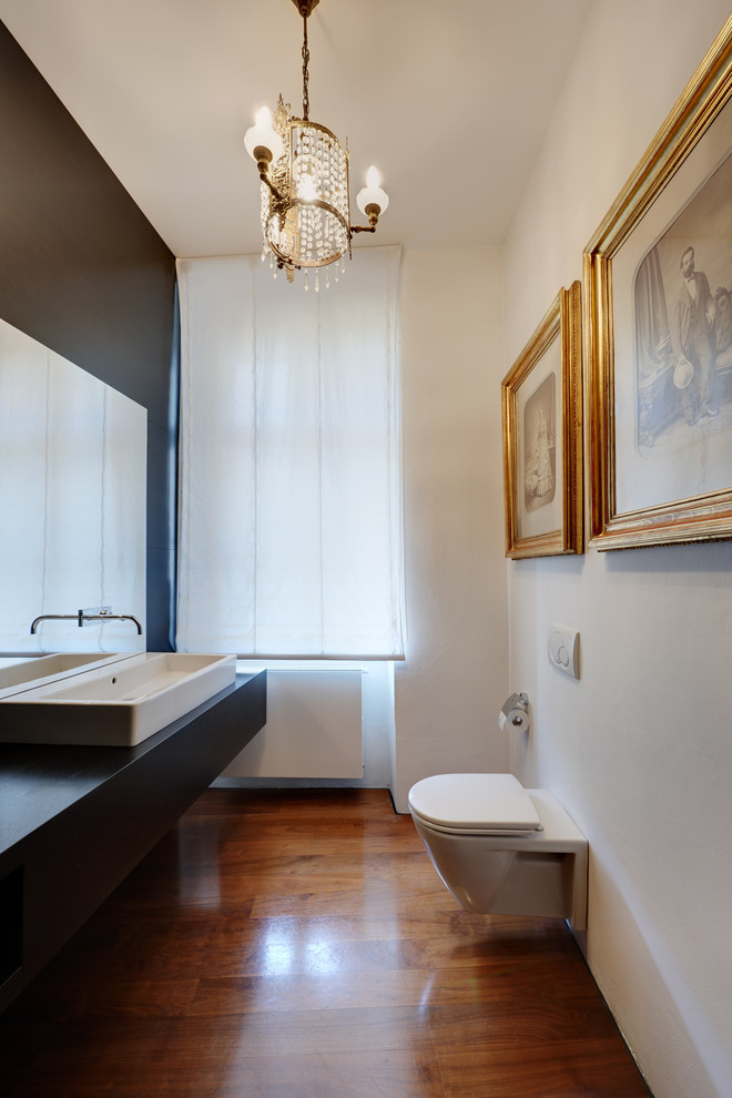 This is an example of a contemporary powder room in Other with a wall-mount toilet, white walls, medium hardwood floors, a vessel sink and black benchtops.