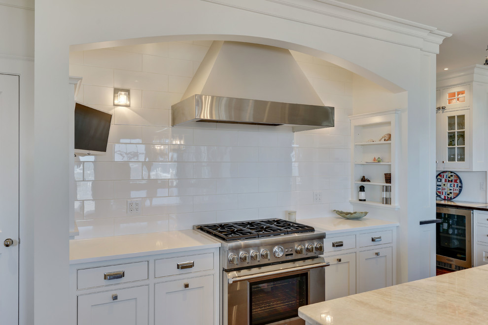 This is an example of an expansive beach style u-shaped kitchen in New York with a drop-in sink, white cabinets, granite benchtops, white splashback, ceramic splashback, stainless steel appliances, dark hardwood floors, with island, brown floor and white benchtop.