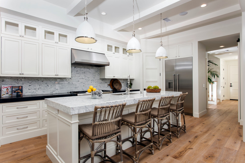 Inspiration for a mid-sized beach style l-shaped open plan kitchen in Orange County with a drop-in sink, shaker cabinets, white cabinets, marble benchtops, white splashback, subway tile splashback, stainless steel appliances, medium hardwood floors and with island.