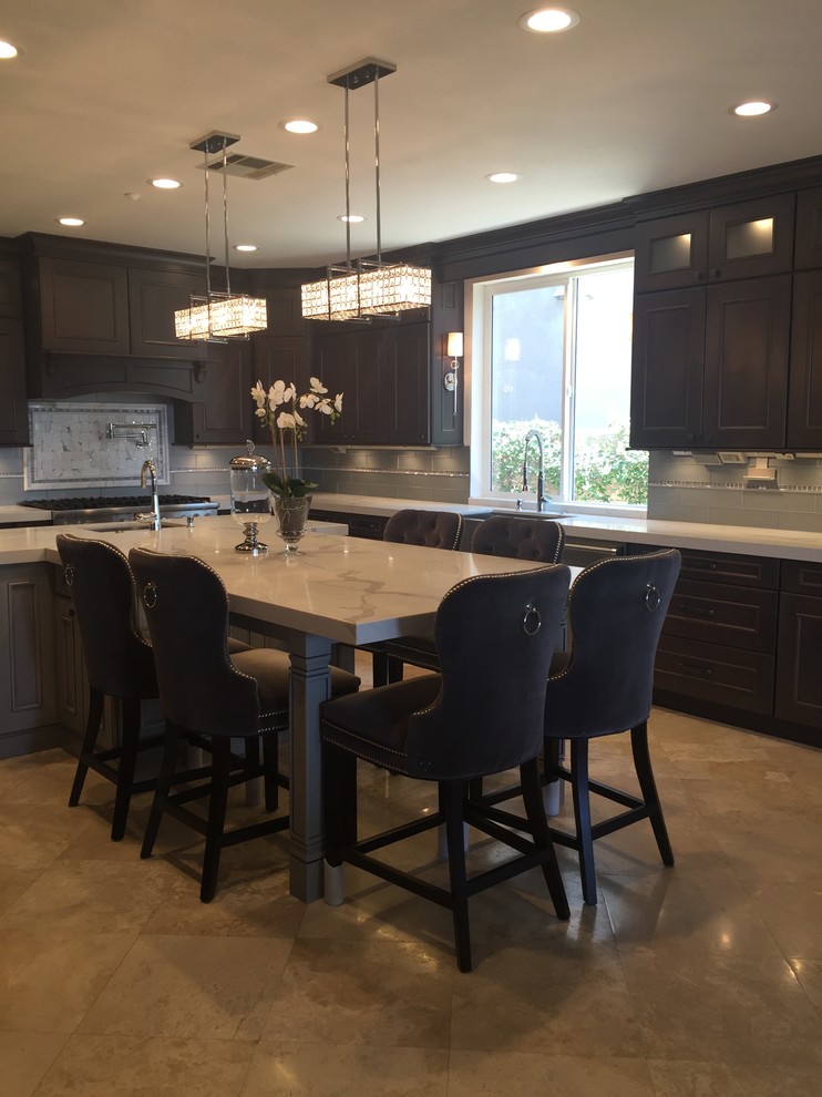 Mid-sized transitional l-shaped travertine floor and beige floor enclosed kitchen photo in Orange County with a farmhouse sink, brown cabinets, marble countertops, gray backsplash, glass tile backsplash, stainless steel appliances, an island and beaded inset cabinets