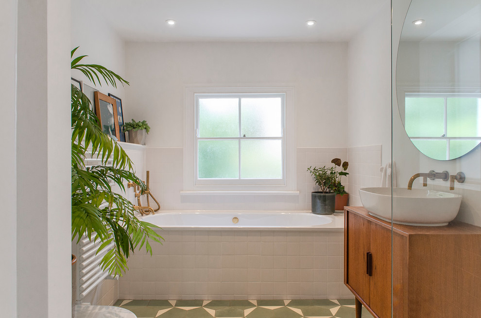 This is an example of a mid-sized modern kids bathroom in London with furniture-like cabinets, medium wood cabinets, a drop-in tub, a corner shower, a wall-mount toilet, white tile, ceramic tile, white walls, cement tiles, a vessel sink, glass benchtops, green floor and an open shower.