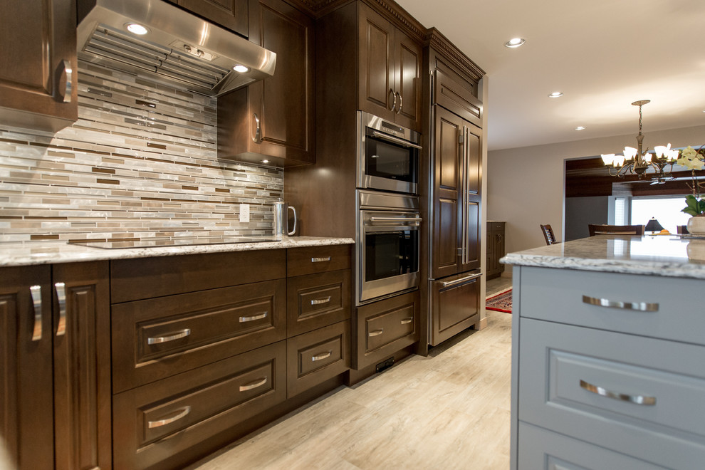 This is an example of a large traditional eat-in kitchen in Vancouver with an undermount sink, raised-panel cabinets, dark wood cabinets, quartz benchtops, grey splashback, glass tile splashback, stainless steel appliances, light hardwood floors and with island.