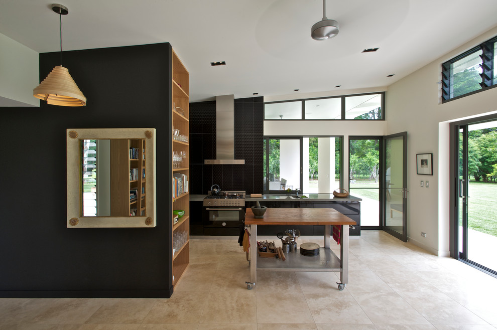 Large contemporary open plan kitchen in Sydney with concrete benchtops, flat-panel cabinets, black splashback, travertine floors, with island, black cabinets and black appliances.