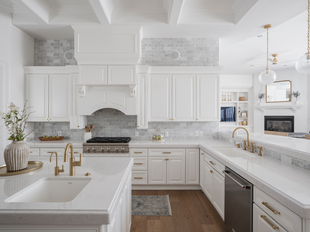 Inredning av ett klassiskt stort vit vitt kök, med en undermonterad diskho, luckor med upphöjd panel, vita skåp, bänkskiva i kvarts, vitt stänkskydd, stänkskydd i marmor, rostfria vitvaror, ljust trägolv, en köksö och beiget golv