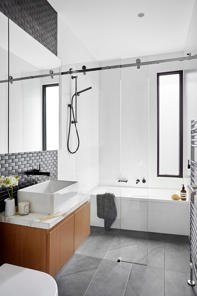 This is an example of a mid-sized contemporary bathroom in Melbourne with flat-panel cabinets, light wood cabinets, a drop-in tub, an alcove shower, a wall-mount toilet, white tile, cement tile, white walls, ceramic floors, a vessel sink, marble benchtops, grey floor and a sliding shower screen.