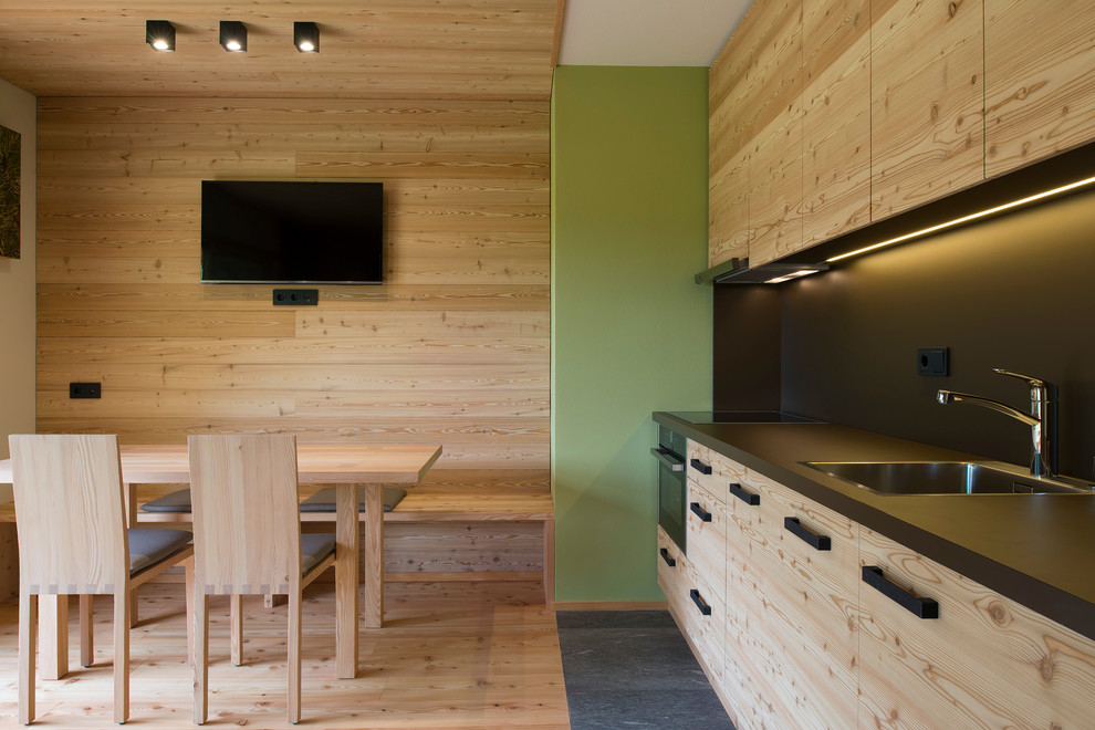 This is an example of a mid-sized country single-wall eat-in kitchen in Other with a drop-in sink, flat-panel cabinets, light wood cabinets, black splashback, black appliances and light hardwood floors.