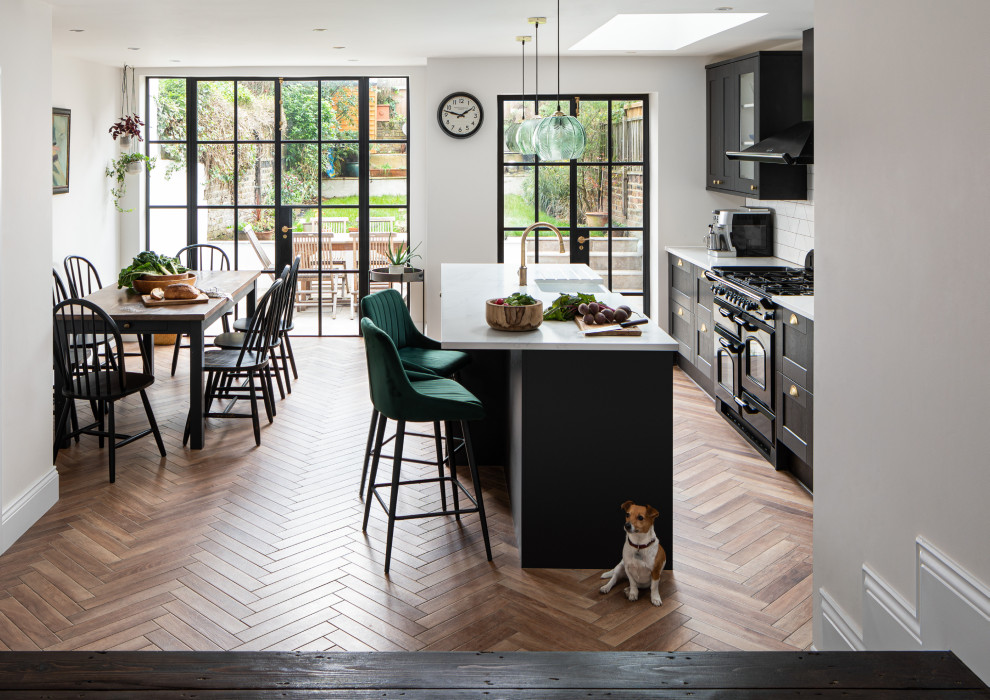 Photo of a transitional galley eat-in kitchen in London with shaker cabinets, black cabinets, white splashback, subway tile splashback, black appliances, medium hardwood floors, with island, brown floor and white benchtop.