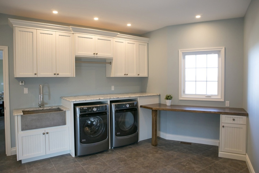Photo of a mid-sized transitional l-shaped utility room in Toronto with a farmhouse sink, shaker cabinets, white cabinets, granite benchtops, grey walls, ceramic floors, a side-by-side washer and dryer and brown floor.