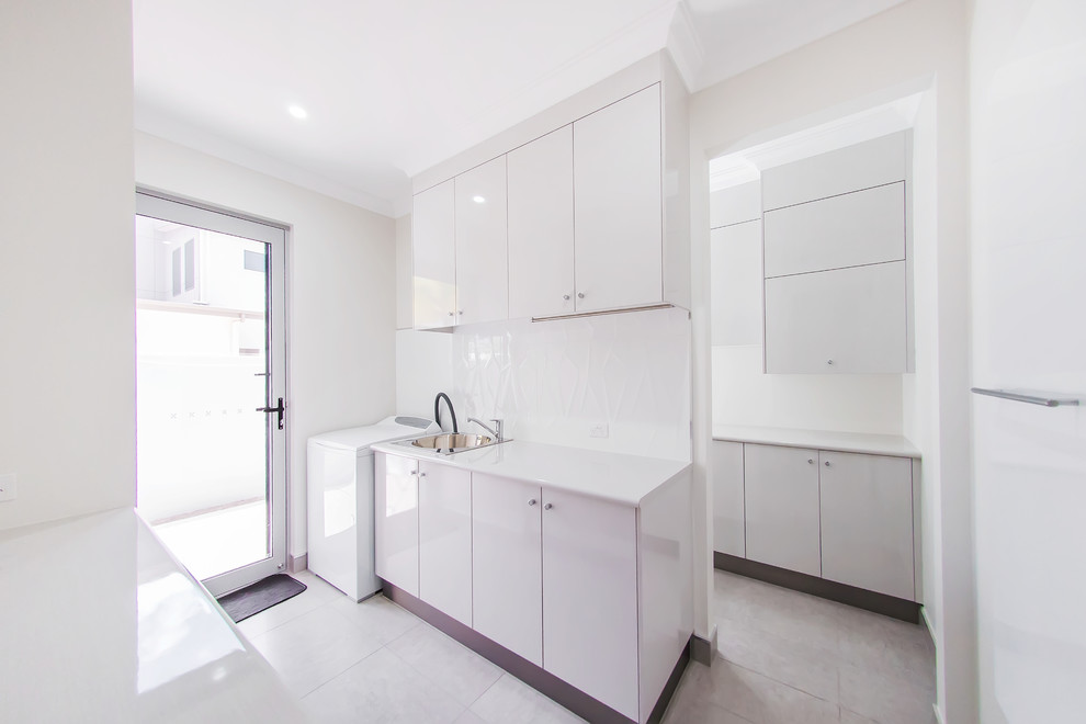 This is an example of a large contemporary galley utility room in Other with a single-bowl sink, flat-panel cabinets, white cabinets, laminate benchtops, white walls, a stacked washer and dryer, white splashback, glass sheet splashback, ceramic floors, grey floor and white benchtop.
