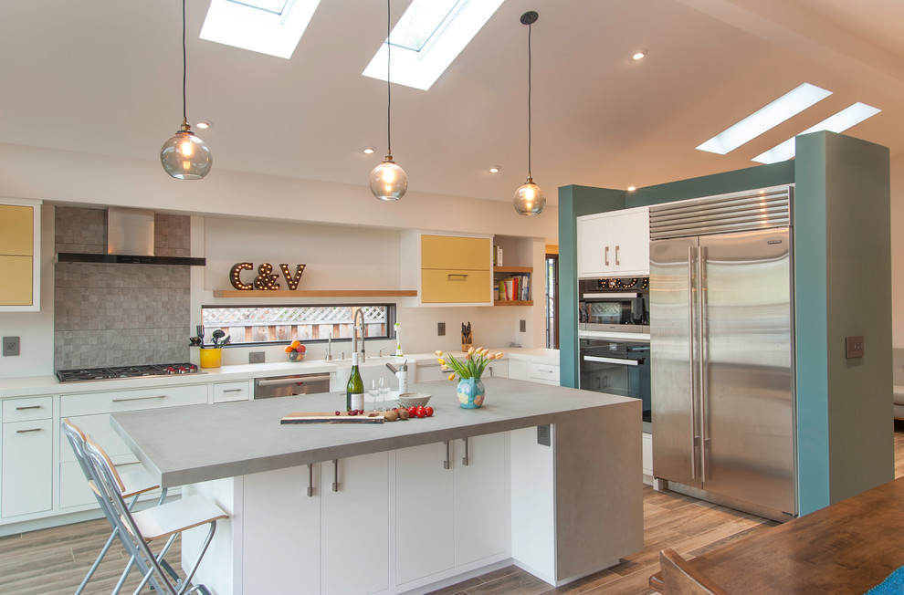 Photo of a mid-sized contemporary l-shaped open plan kitchen in San Francisco with a farmhouse sink, flat-panel cabinets, yellow cabinets, concrete benchtops, grey splashback, cement tile splashback, stainless steel appliances, porcelain floors and with island.