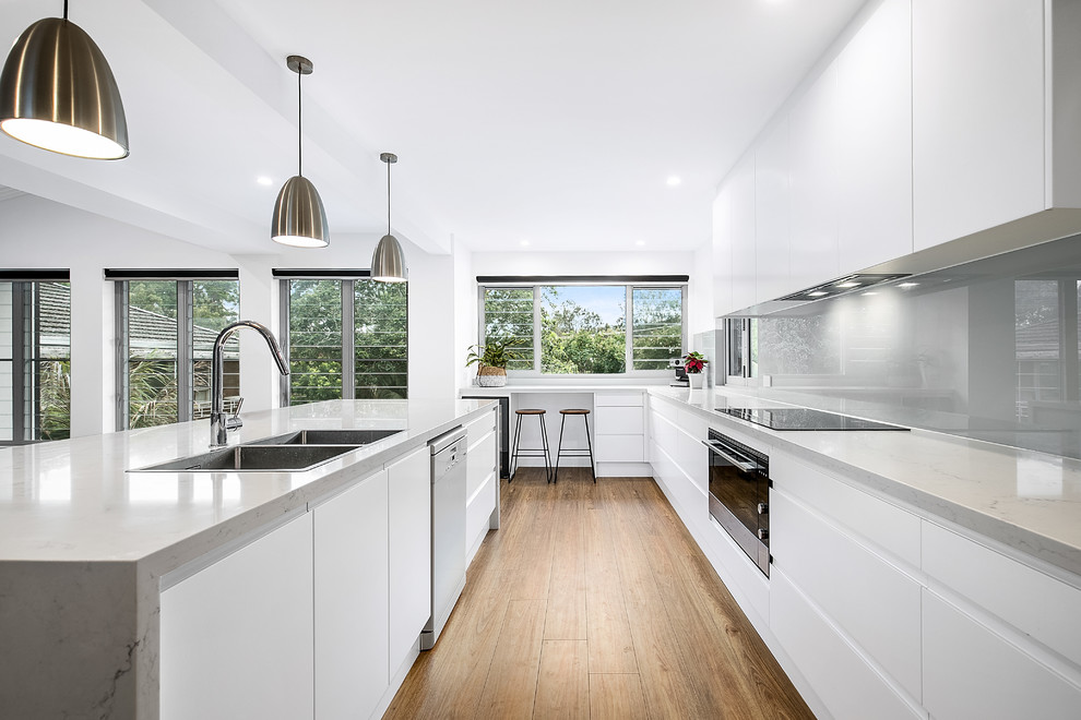 This is an example of a large contemporary l-shaped eat-in kitchen in Sunshine Coast with an undermount sink, flat-panel cabinets, white cabinets, quartz benchtops, grey splashback, glass sheet splashback, stainless steel appliances, dark hardwood floors, with island, brown floor and grey benchtop.