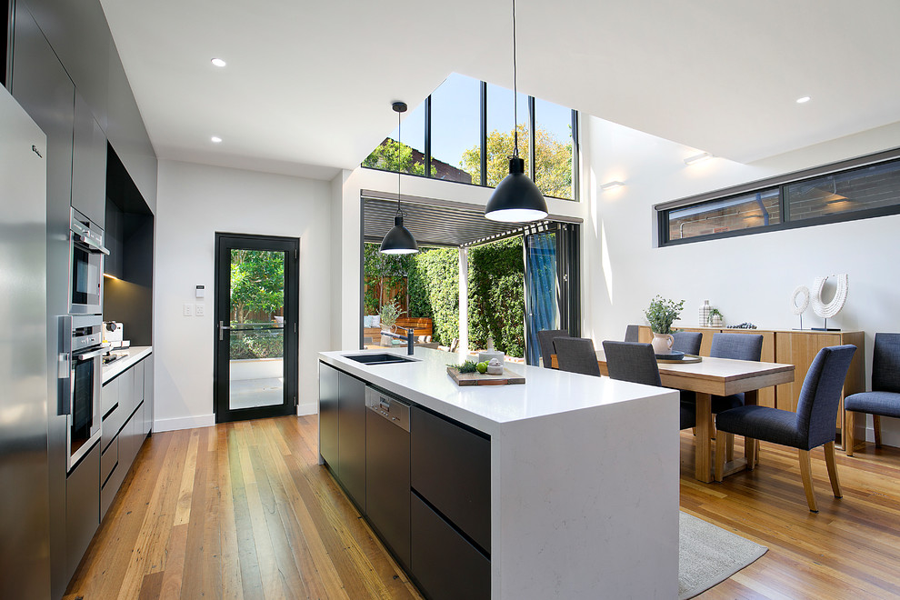 Photo of a contemporary galley open plan kitchen in Sydney with an undermount sink, flat-panel cabinets, black cabinets, marble benchtops, stainless steel appliances, medium hardwood floors, with island, brown floor and white benchtop.