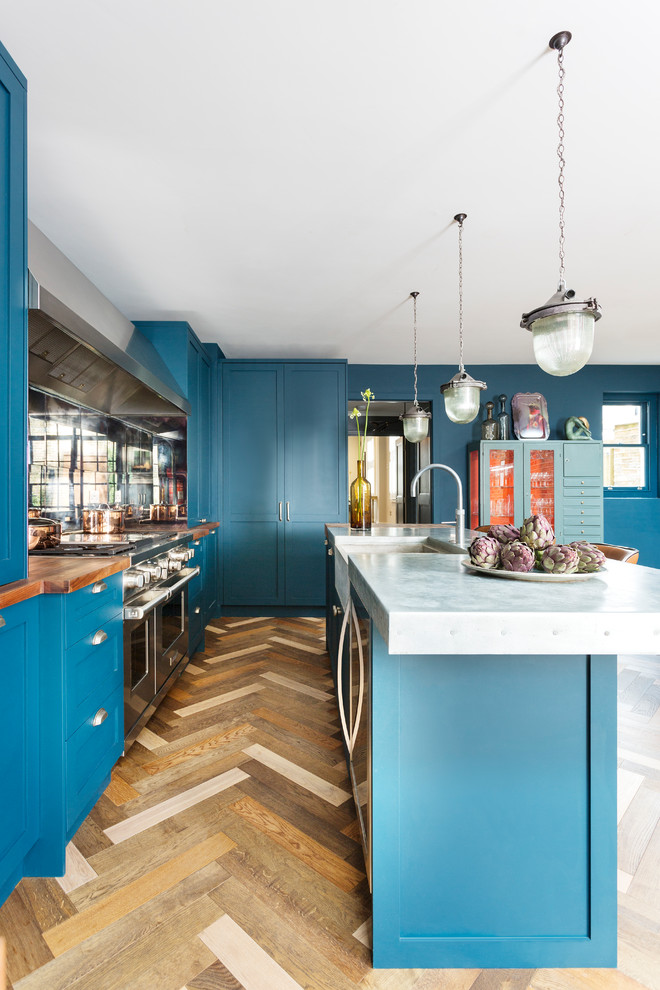 Mid-sized eclectic l-shaped open plan kitchen in London with a farmhouse sink, shaker cabinets, blue cabinets, zinc benchtops, mirror splashback, stainless steel appliances, dark hardwood floors, with island, multi-coloured floor and grey benchtop.