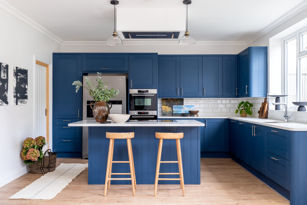 This is an example of a transitional u-shaped kitchen in London with an undermount sink, shaker cabinets, blue cabinets, white splashback, subway tile splashback, stainless steel appliances, light hardwood floors, with island, beige floor and white benchtop.